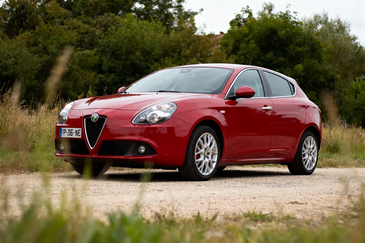 Voiture rouge Alfa Romeo sur chemin gravillonné entouré d'herbe et d'arbres.