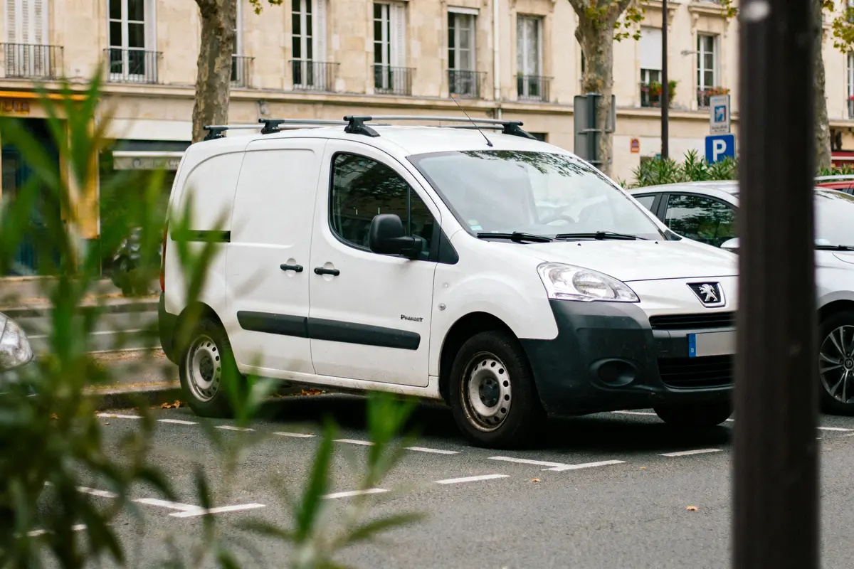 Fourgonnette blanche Peugeot garée en ville, carrefour, arbres, façades d’immeubles, plantes au premier plan.