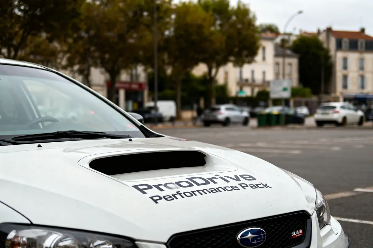 Capot d'une voiture blanche "ProDrive Performance Pack" garée dans une rue avec bâtiments en arrière-plan.