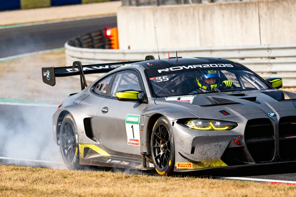 Voiture de course BMW avec pneus fumants sur circuit, pilote concentré, livrée grise et jaune.