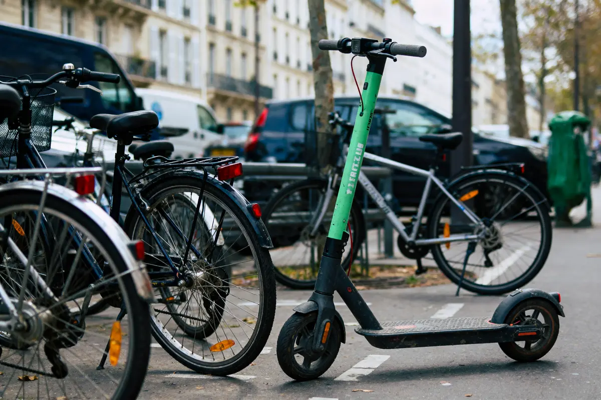 Trottinette électrique verte et vélos stationnés sur un trottoir urbain, voitures en arrière-plan.