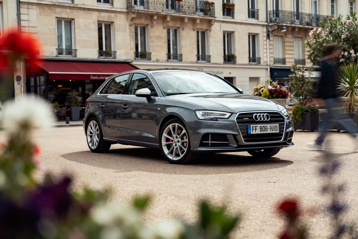 Voiture Audi grise stationnée devant un café parisien, fleurs floues au premier plan, passant en mouvement.