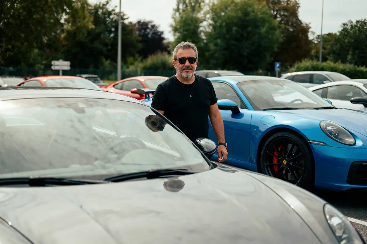 Homme en t-shirt noir devant des voitures de sport, dont une bleue et une grise, stationnées à l'extérieur.
