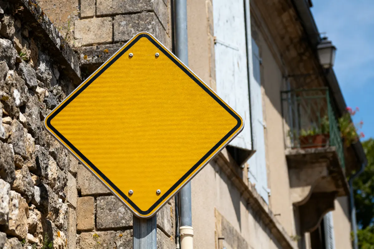 Panneau losange jaune devant un mur en pierre et une maison ancienne